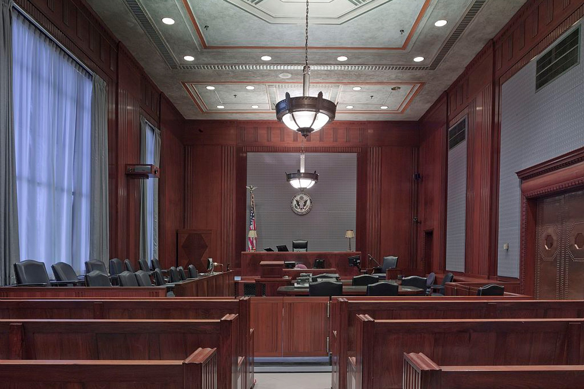 An empty courtroom with wooden walls and benches, and an ornate light fixture hanging from the center.