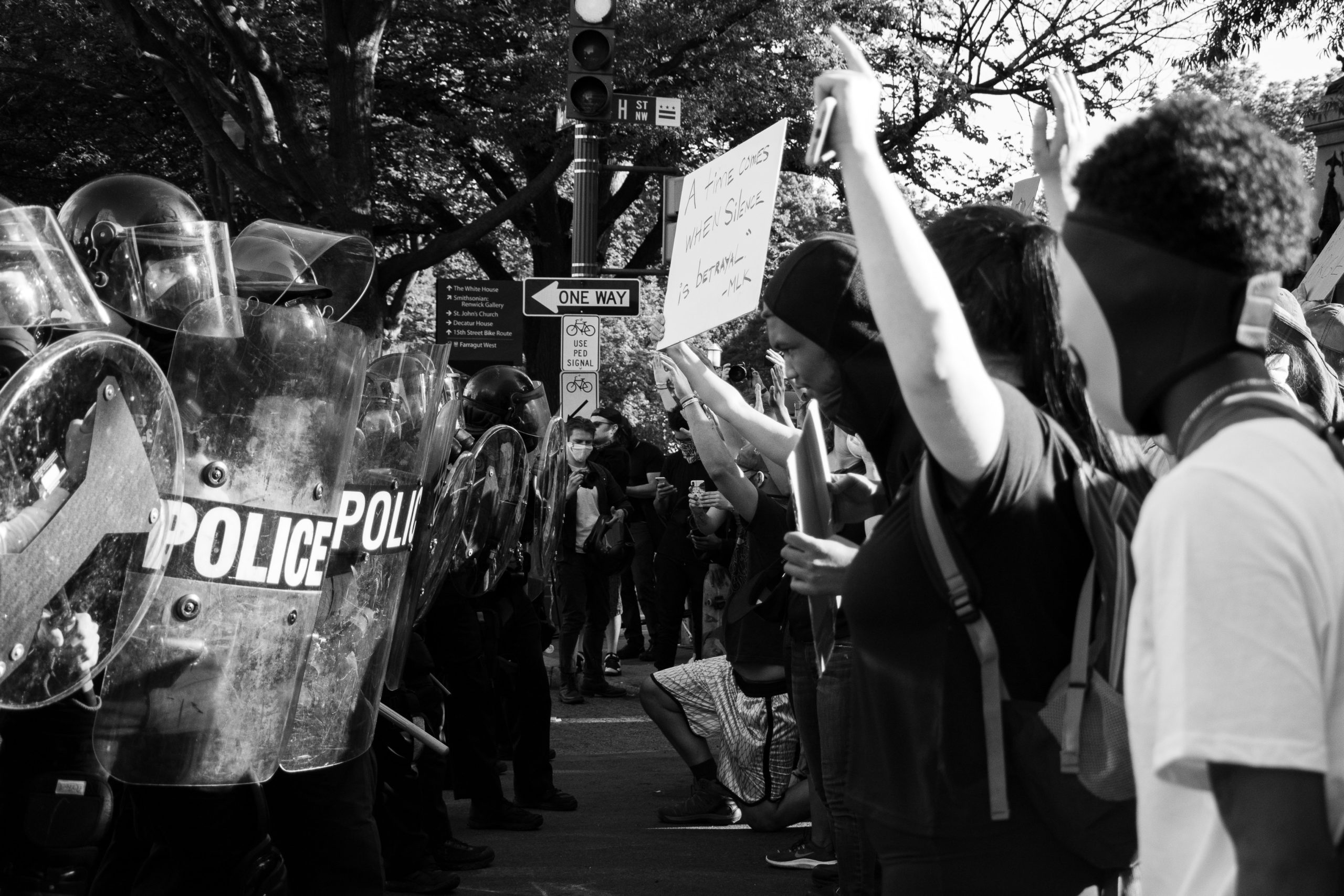 To the left of the black and white image, there is a row of police in gear and facing across from them, there is a row of protesters with signs.