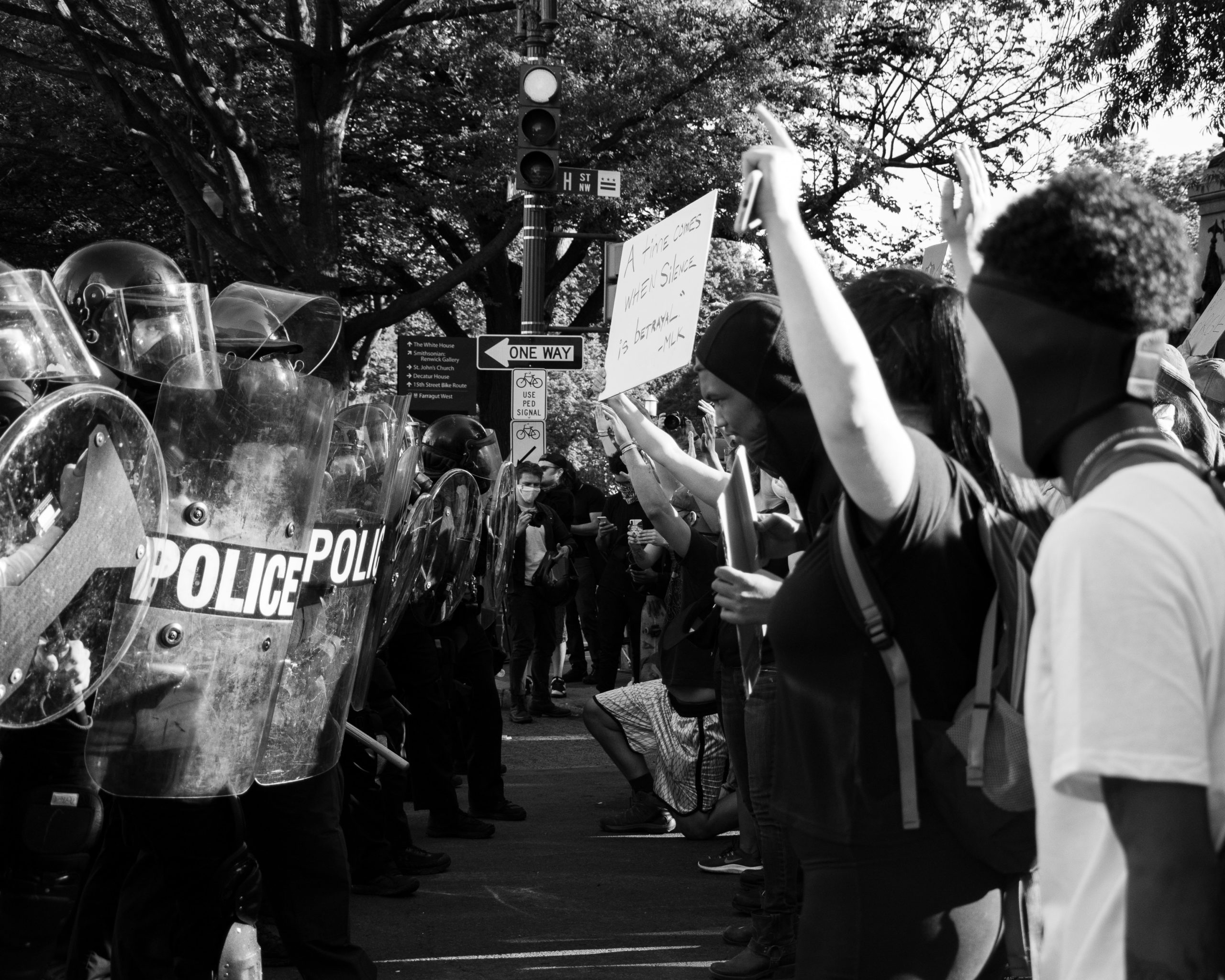 To the left of the black and white image, there is a row of police in gear and facing across from them, there is a row of protesters with signs.
